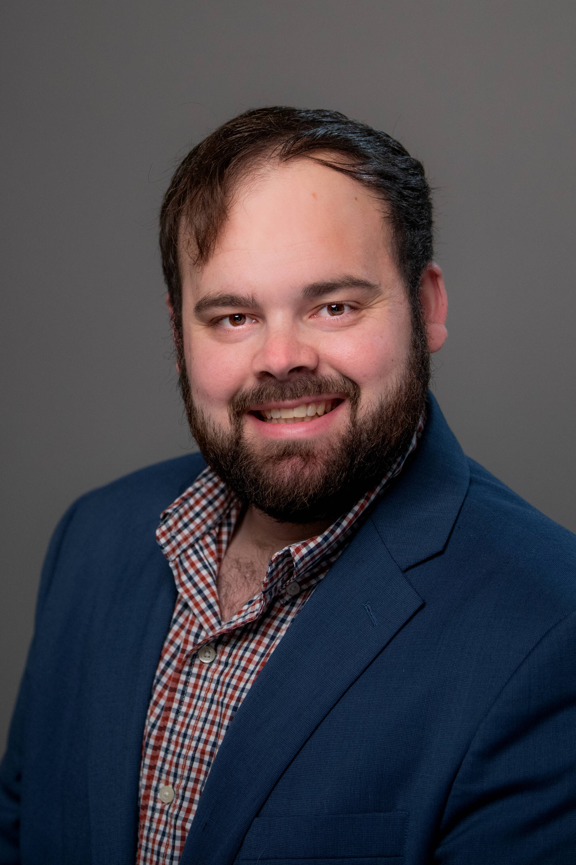 Brock Thane in a navy blazer with a plaid shirt on underneath smiling at the camera.